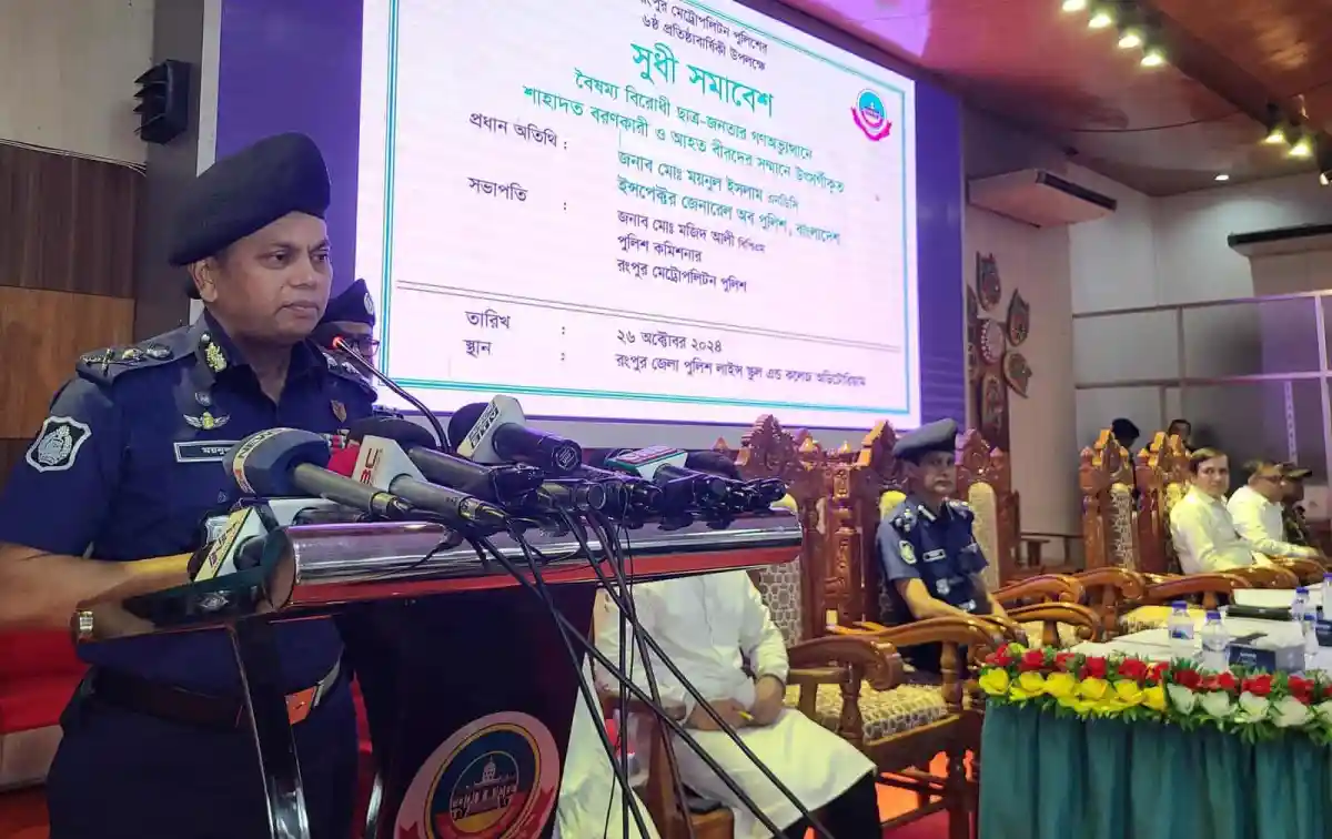 Bangladesh police officer speaking at a formal event with microphones and a large screen behind. Channel i News - চ্যানেল আই নিউজ