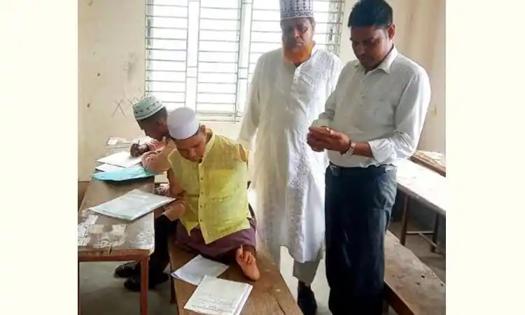 রাসেল মৃধা পা দিয়ে লিখে আলিম পাস করছেন, পাশে দুই ব্যক্তি দেখছেন. Channel i News - চ্যানেল আই নিউজ