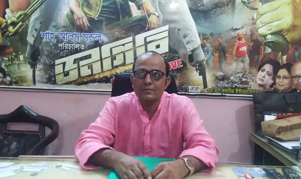 বাংলাদেশের একজন সংবাদ প্রতিবেদক বসে আছেন, পেছনে সিনেমার পোস্টার. Channel i News - চ্যানেল আই নিউজ