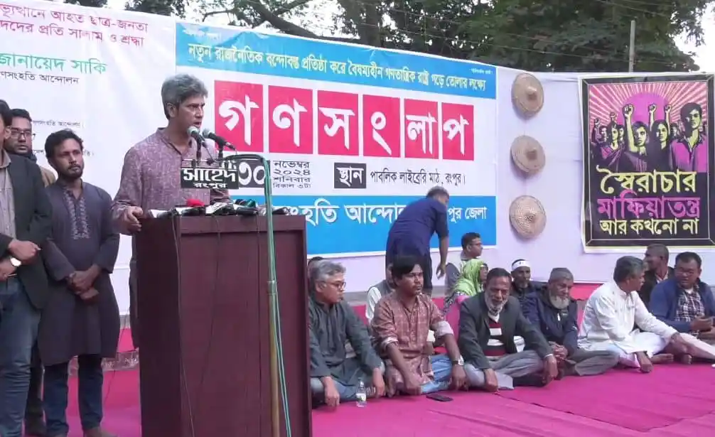 বক্তব্য দিচ্ছেন একজন বক্তা, পাশে বসে আছেন অন্যরা, ব্যানারে লেখা গানের শপ ও বিভিন্ন ছবি. Channel i News - চ্যানেল আই নিউজ