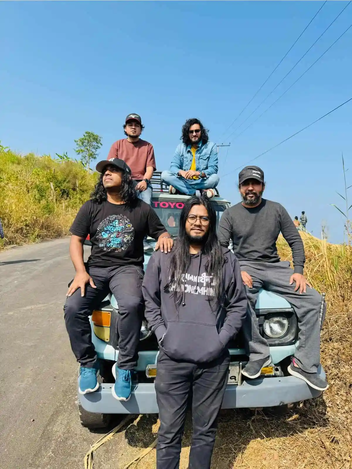 Shironamhin band members posing on a rural road with a car and greenery. Channel i News - চ্যানেল আই নিউজ