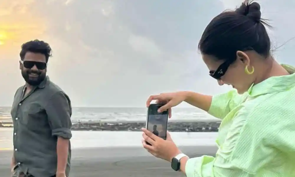নারী স্মার্টফোন দিয়ে ছবি তুলছে, পেছনে পুরুষ হাসছে. Channel i News - চ্যানেল আই নিউজ