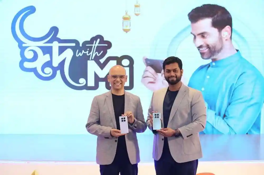 দুই ব্যক্তি মোবাইল ফোন দেখাচ্ছেন, পেছনে চ্যানেল আই লোগো. Channel i News - চ্যানেল আই নিউজ