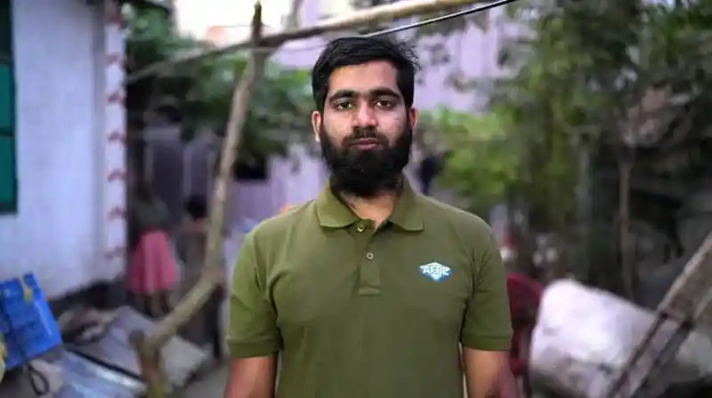 বাংলাদেশি যুবক গাছের নিচে দাঁড়িয়ে, পটভূমিতে বাড়ি ও গাছের দৃশ্য. Channel i News - চ্যানেল আই নিউজ