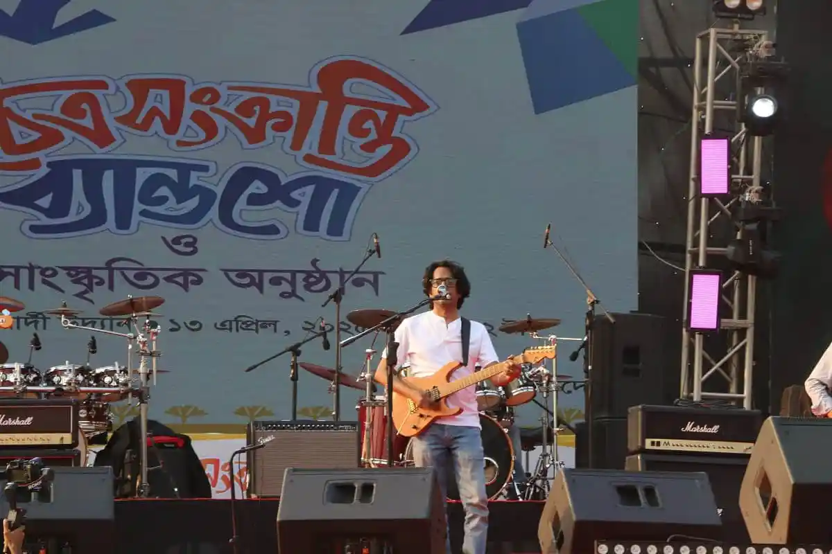 বৃহৎ কনসার্টে গিটার বাজাচ্ছেন একজন শিল্পী, পেছনে ব্যান্ড সদস্যরা ও বড় স্ক্রিনে বাংলা লেখা. Channel i News - চ্যানেল আই নিউজ