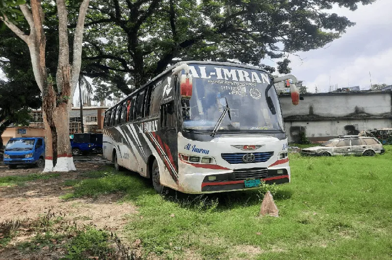 বাসটি একটি খোলা মাঠে দাঁড়িয়ে আছে, চারপাশে গাছ ও অন্যান্য যানবাহন. Channel i News - চ্যানেল আই নিউজ