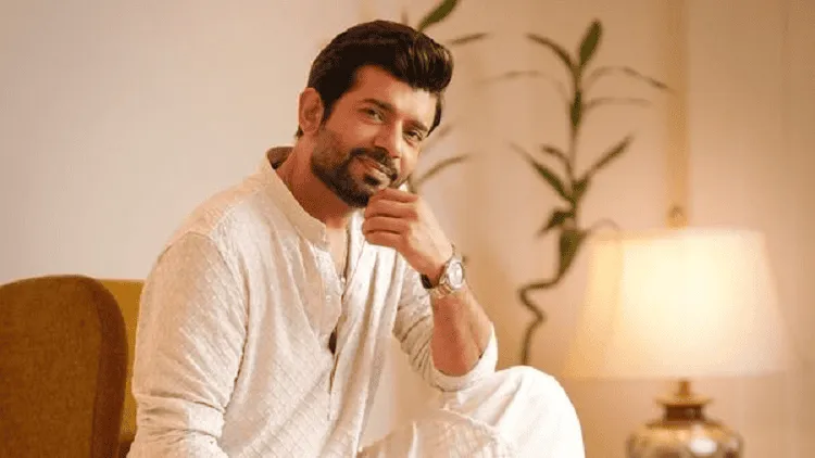 অভিনেতা হাস্যোজ্জ্বল মুখে বসে আছেন, পেছনে ল্যাম্প ও গাছের পাতা. Channel i News - চ্যানেল আই নিউজ