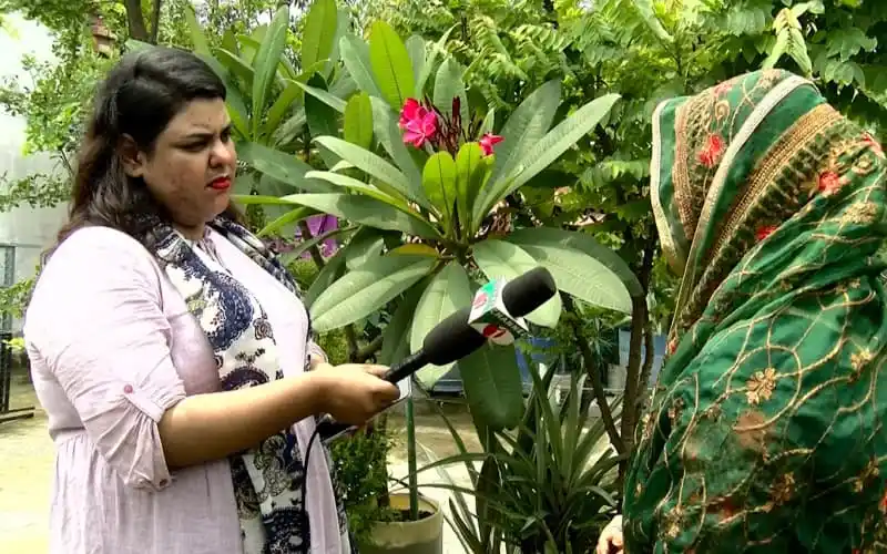 একজন নারী সাংবাদিক ফুলের গাছের সামনে সাক্ষাৎকার নিচ্ছেন. Channel i News - চ্যানেল আই নিউজ