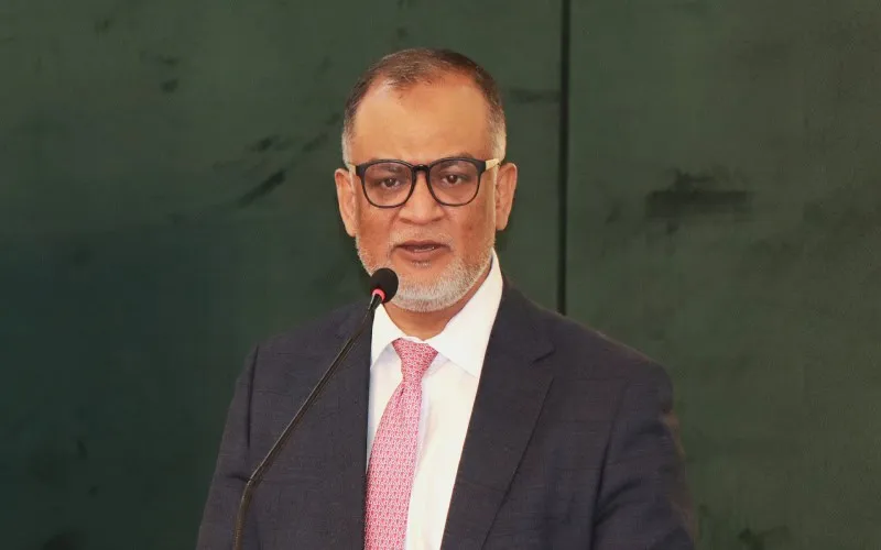 বাংলাদেশের বাণিজ্য উপদেষ্টা বক্তৃতা দিচ্ছেন, পেছনে সবুজ পটভূমি. Channel i News - চ্যানেল আই নিউজ