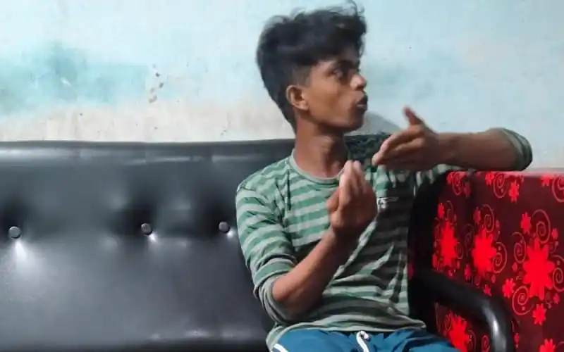 Young boy sitting on a black sofa, gesturing with his hands, in a room with a light blue wall. Channel i News - চ্যানেল আই নিউজ