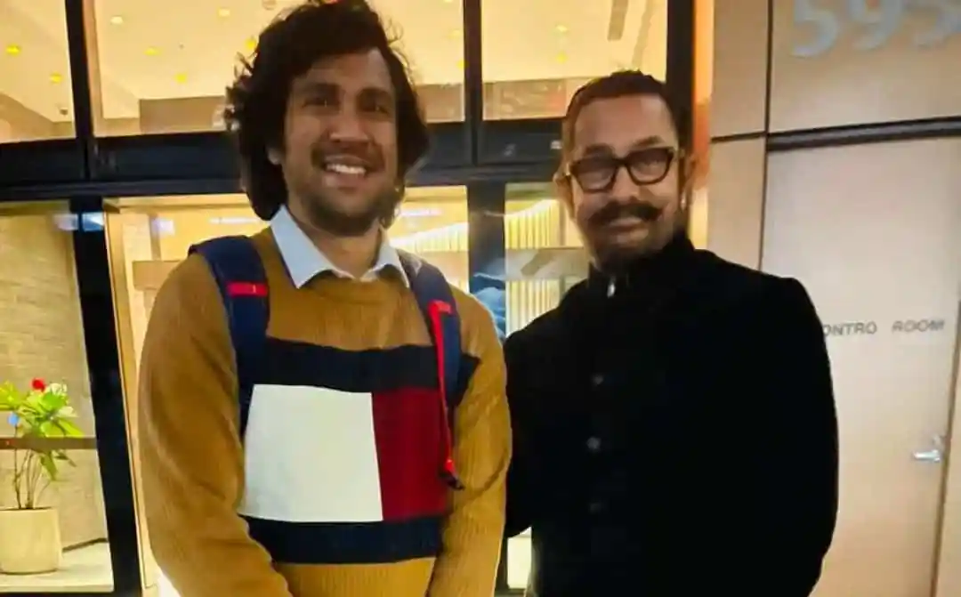 দুই ব্যক্তি হাসিমুখে একসঙ্গে দাঁড়িয়ে, পেছনে আধুনিক ভবনের জানালা।. Channel i News - চ্যানেল আই নিউজ