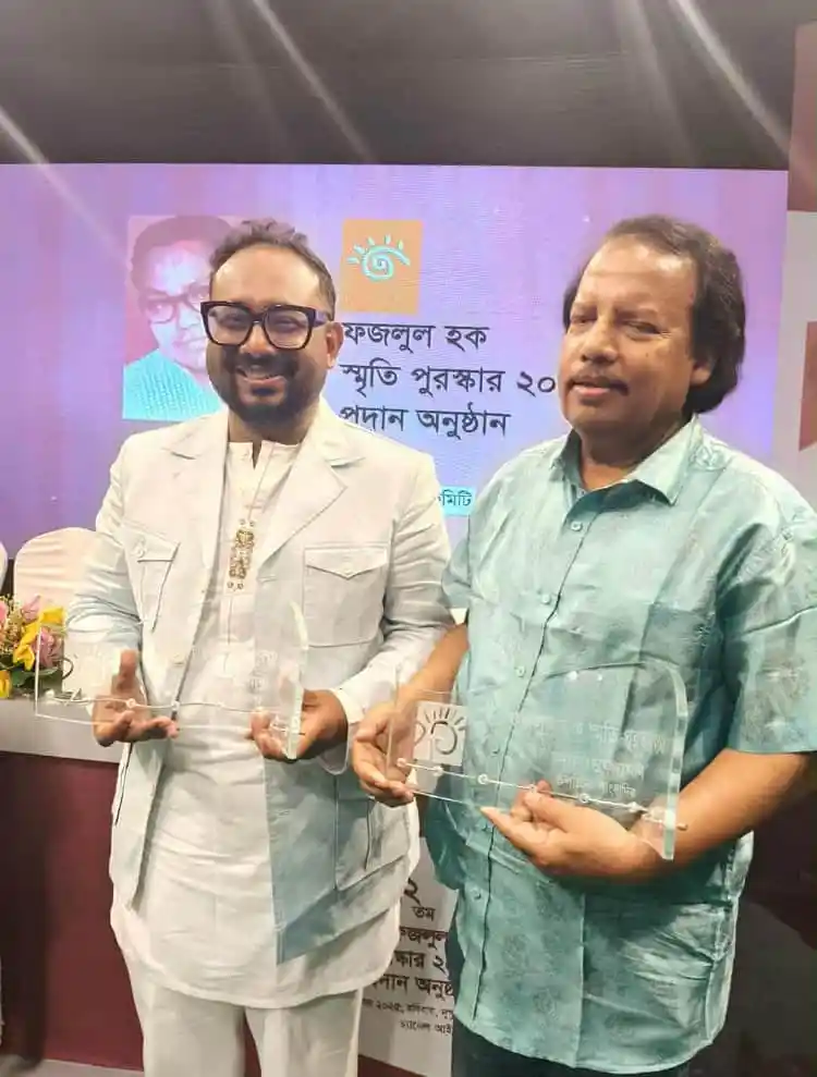 দুই ব্যক্তি সম্মাননা পুরস্কার গ্রহণ করছেন, পেছনে ব্যানার দেখা যাচ্ছে. Channel i News - চ্যানেল আই নিউজ