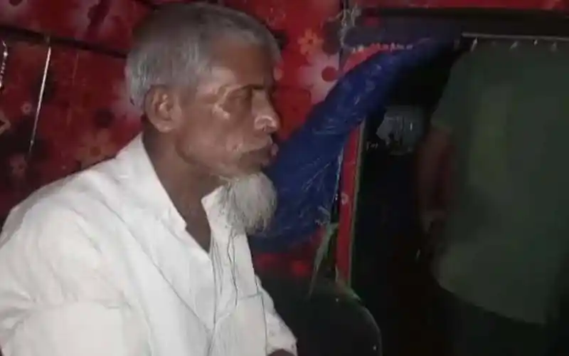 Older man with white beard and white shirt sitting indoors in a dimly lit room. Channel i News - চ্যানেল আই নিউজ