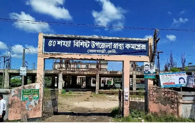 Feni district hospital entrance with blue sky and construction site in background. Channel i News - চ্যানেল আই নিউজ