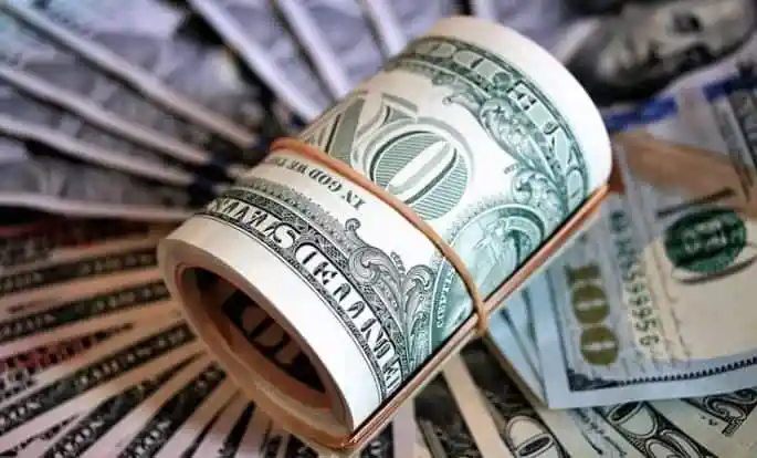 Roll of US dollar bills with a rubber band on top of scattered dollar notes. Channel i News - চ্যানেল আই নিউজ