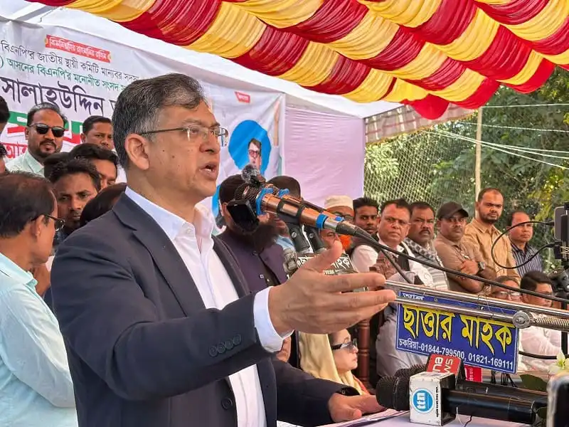 Bangladeshi politician speaking at a public event with a crowd and banners. Channel i News - চ্যানেল আই নিউজ