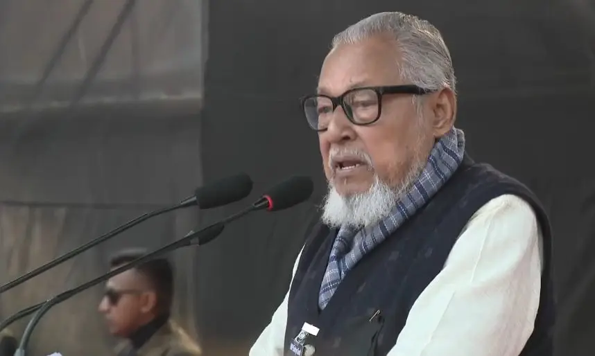 বয়স্ক ব্যক্তি ভাষণ দিচ্ছেন, পেছনে অন্য ব্যক্তি দেখা যাচ্ছে. Channel i News - চ্যানেল আই নিউজ