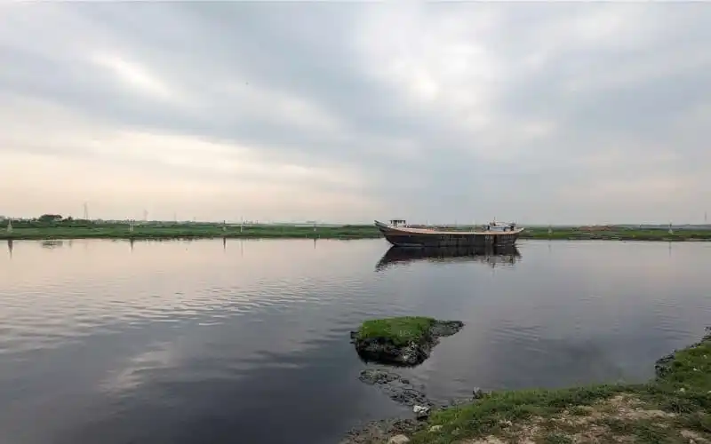 নদীতে একটি নৌকা ভাসছে, আকাশ আংশিক মেঘলা, শান্ত জলরাশি. Channel i News - চ্যানেল আই নিউজ
