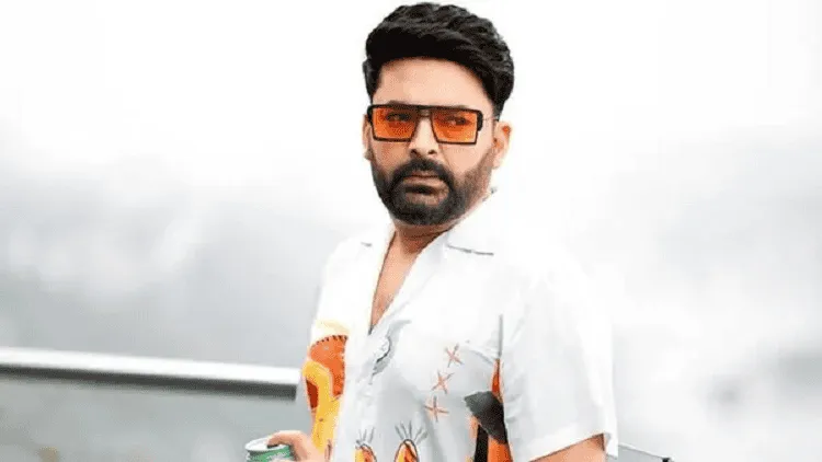 কপিল শর্মা ক্যামেরার সামনে দাঁড়িয়ে আছেন, সাদা শার্ট পরা. Channel i News - চ্যানেল আই নিউজ