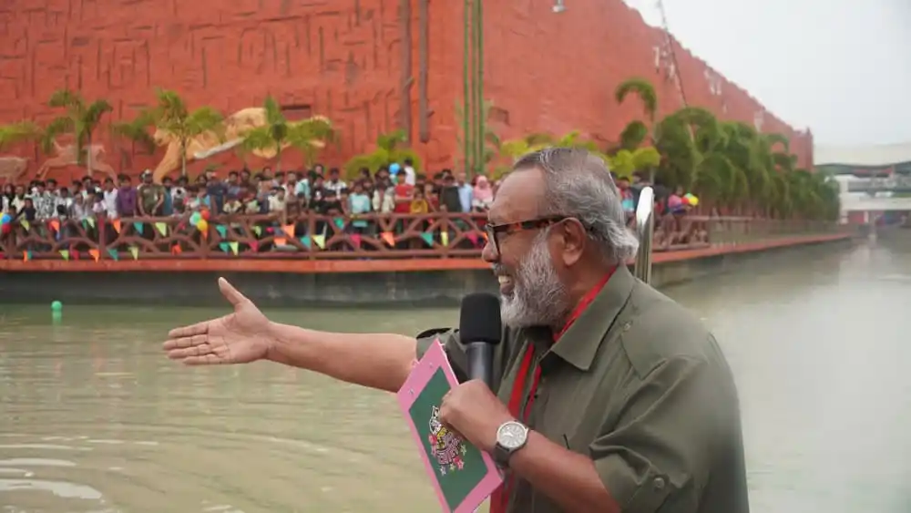 একজন সাংবাদিক নদীর পাড়ে বক্তব্য দিচ্ছেন, পেছনে দর্শকদের ভিড়. Channel i News - চ্যানেল আই নিউজ
