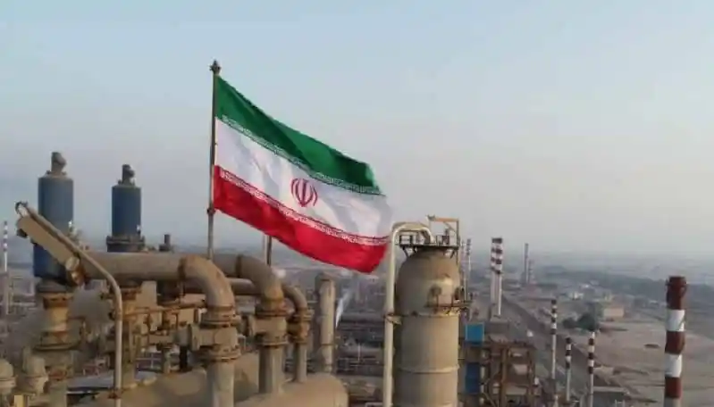 Iran flag flying over industrial refinery site with pipes and smokestacks. Channel i News - চ্যানেল আই নিউজ