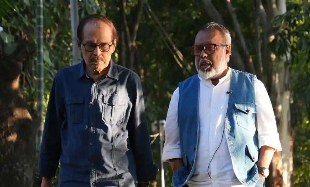 দুই ব্যক্তি বনজঙ্গলে হাঁটছেন, একজনের চোখে চশমা, অন্যজনের গেঞ্জি ও জ্যাকেট পরা. Channel i News - চ্যানেল আই নিউজ
