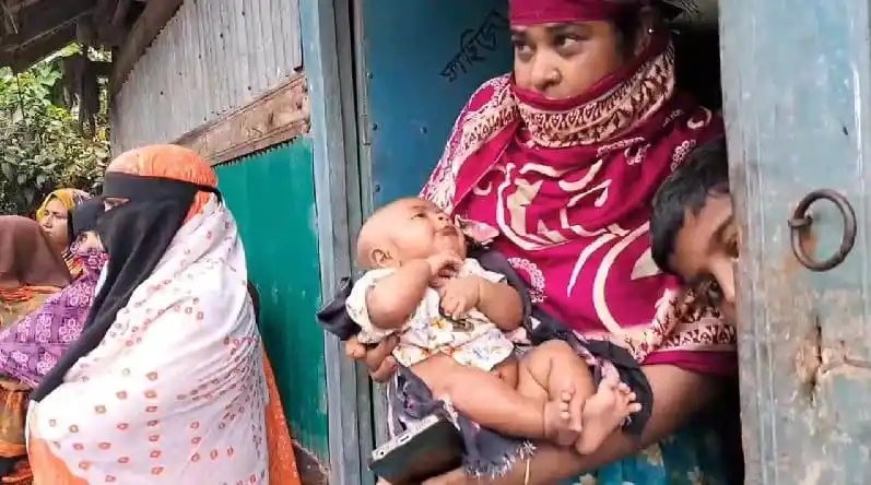 একজন নারী শিশুকে কোলে নিয়ে উদ্বিগ্নভাবে দেখছেন, অন্যরা পাশে দাঁড়িয়ে।. Channel i News - চ্যানেল আই নিউজ