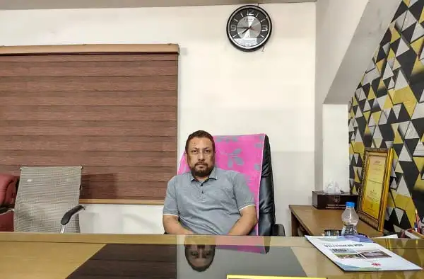 বাংলাদেশি ব্যক্তি অফিসে বসে আছেন, পেছনে ঘড়ি ও ডেস্কের বিভিন্ন সামগ্রী. Channel i News - চ্যানেল আই নিউজ