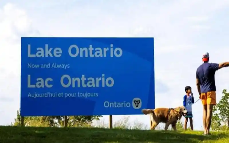 Lake Ontario sign with people and dog in foreground, clear sky background. Channel i News - চ্যানেল আই নিউজ